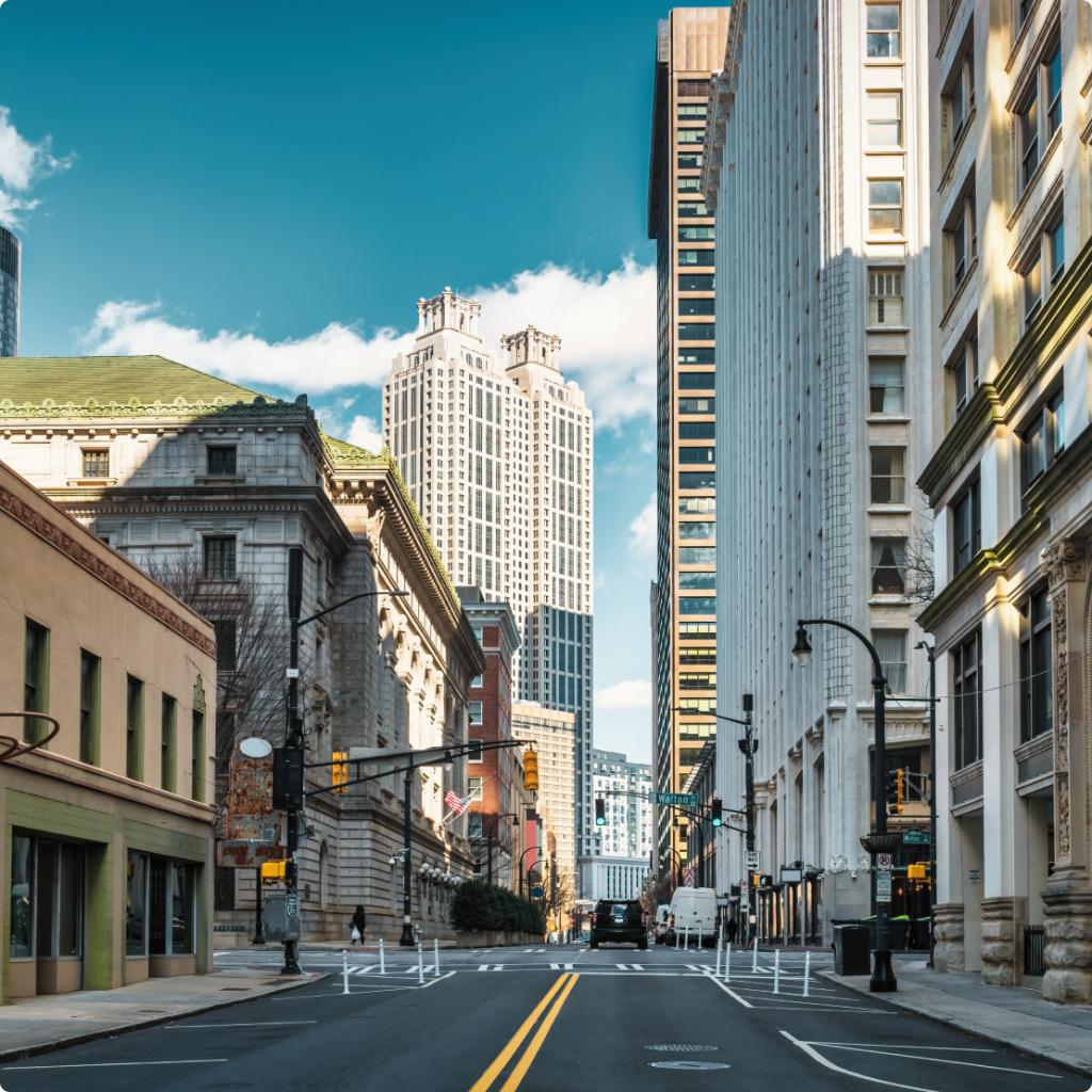 A city street flanked by tall modern and historic buildings under a clear blue sky.
