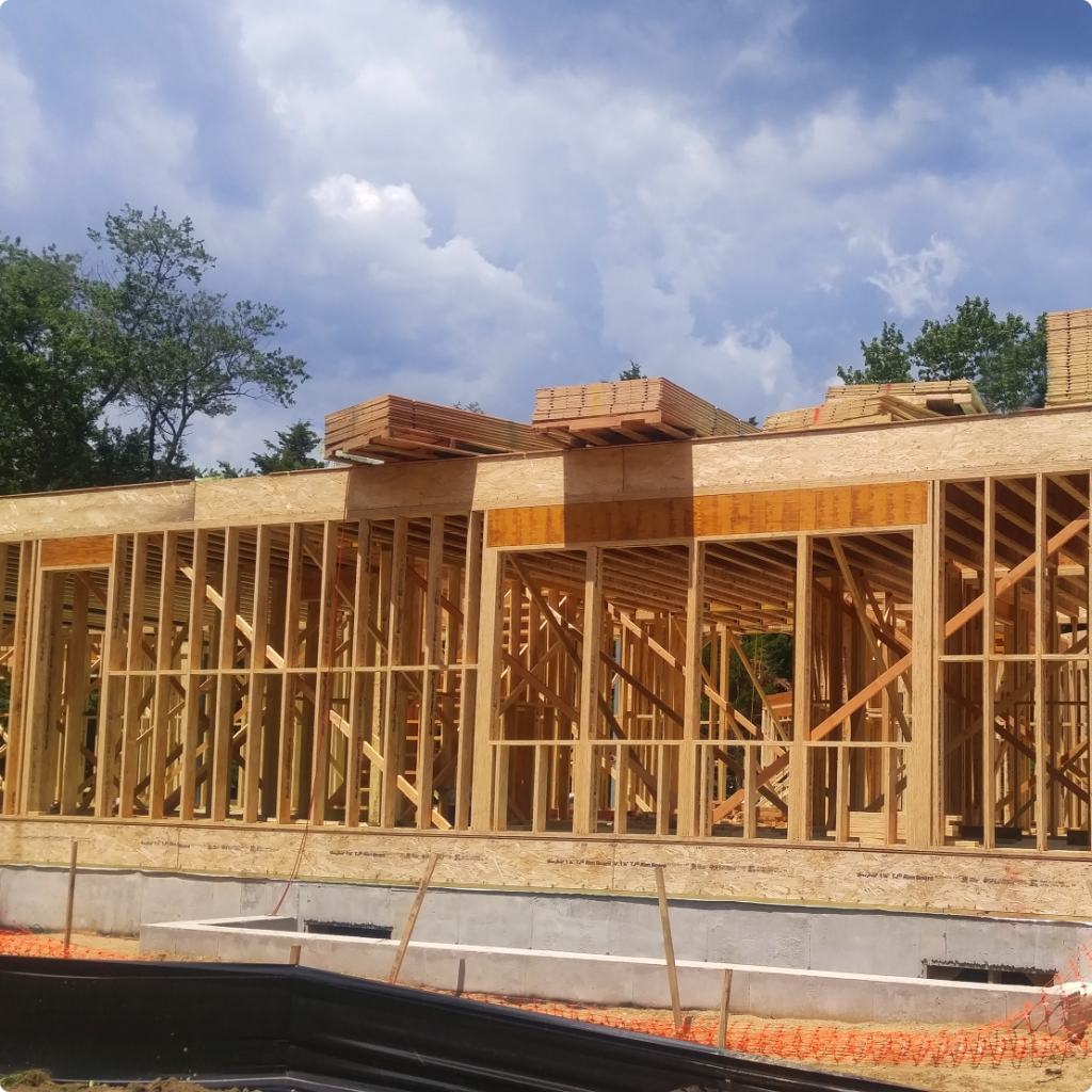 A wooden-framed building under construction with exposed studs and stacks of lumber on top, set on a concrete foundation under a blue sky.