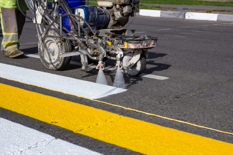 A road line striping machine paints white lane lines on a paved street beside yellow road markings.