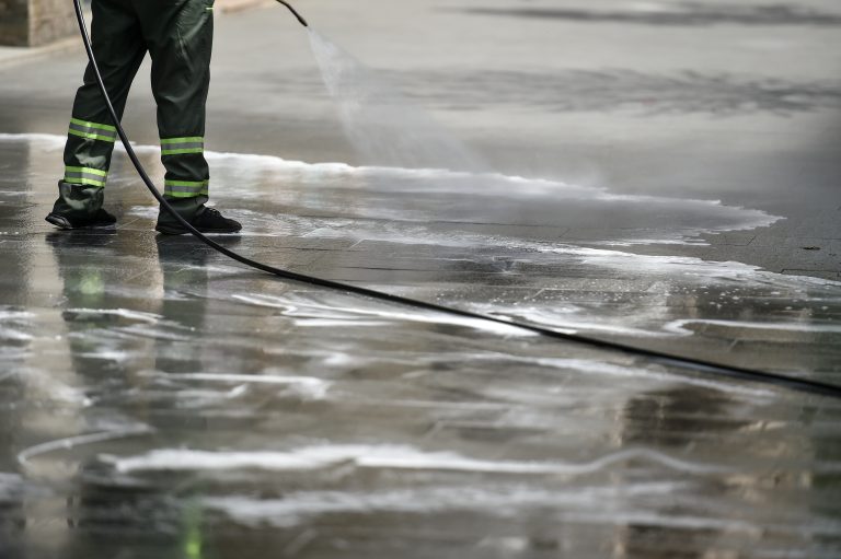 A person in dark pants and boots uses a hose to spray water on a wet street.