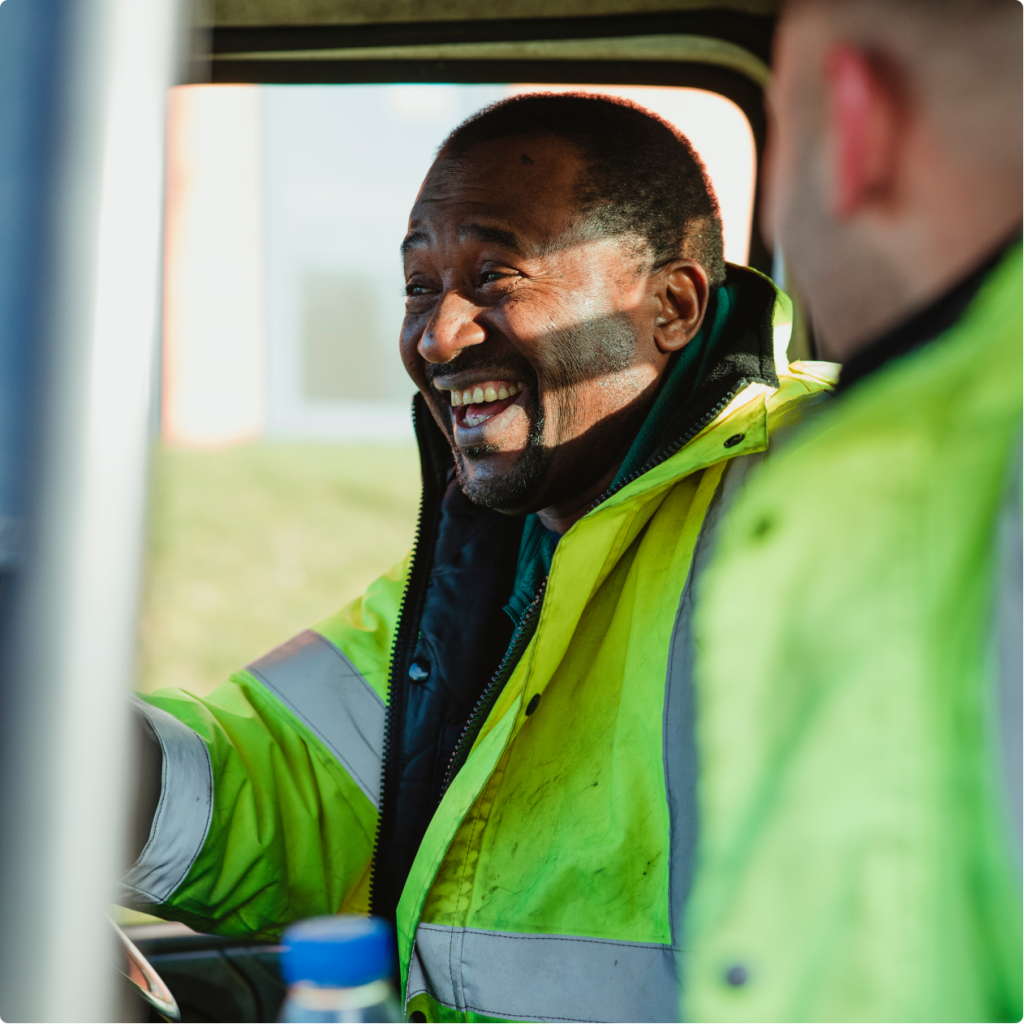 A smiling man in a neon high-visibility jacket sits in the cab of a vehicle with a colleague wearing a similar jacket.