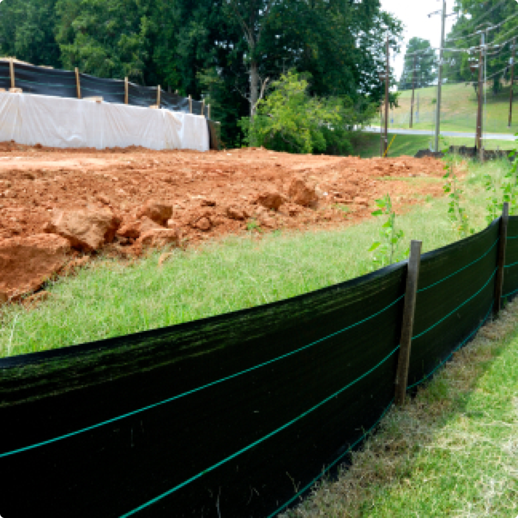 A construction site with a mound of orange soil and a black silt fence along the grassy foreground.