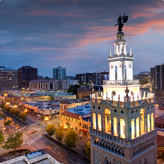 Tall ornate tower topped with a statue rises above a city at dusk, with illuminated streets and buildings.
