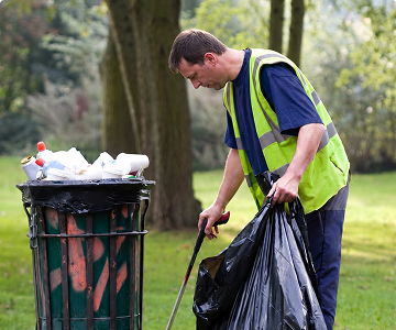 A man in a neon safety vest picks up litter in a park with a grabber, placing trash into a black bag beside a green trash can.