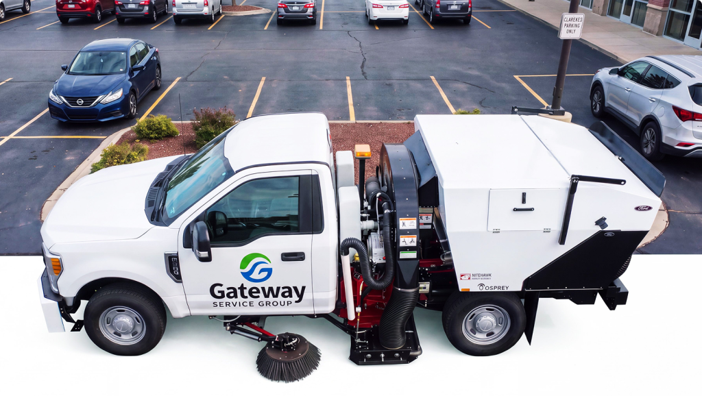 White Gateway lot sweeping truck with a large enclosed back compartment, a side ladder, and Gateway branding, parked in a parking lot near a manhole cover.