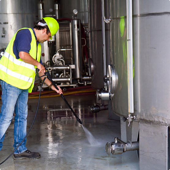 A worker in a yellow safety vest and hard hat uses a pressure washer to clean the floor beside industrial equipment.